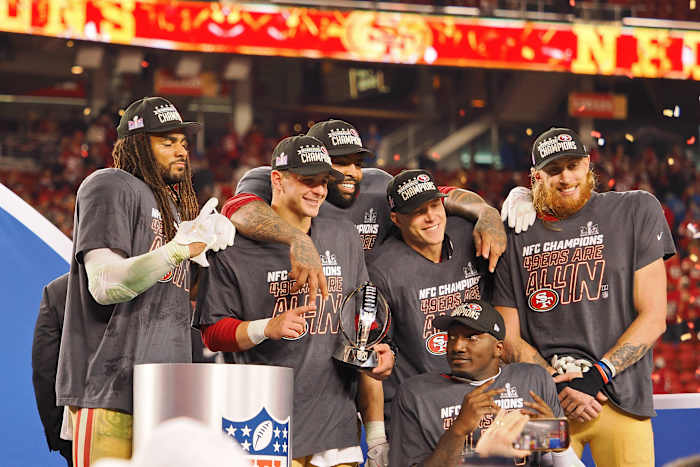 Fred Warner (54), quarterback Brock Purdy (13), offensive tackle Trent Williams (71), running back Christian McCaffrey (23), wide receiver Deebo Samuel (19), and tight end George Kittle all stand in a group smiling after winning